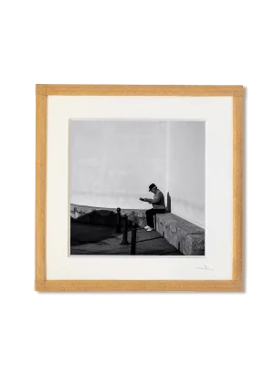 tirage photo petit format encadré à la main représentant un vieil homme assis contre un mur lisant le journal
