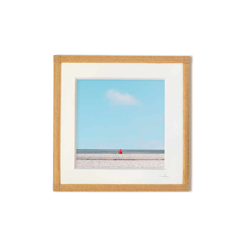 tirage photo petit format encadré à la main représentant un petit personnage rouge assis face à l'océan avec ciel bleu et un nuage